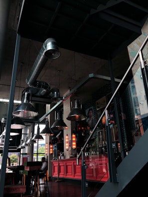 A modern, industrial-style interior with exposed ductwork and large, metallic pendant lights hanging from the ceiling. There is a red-painted bar or counter area, with vertical neon lights illuminating the space. A few tables and chairs are visible, and the area is decorated with various artworks on the wall.
