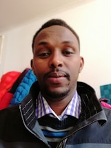 A person wearing a multi-layered outfit with a checked shirt, striped sweater, and dark jacket. The background is a plain wall with clothing items hanging.