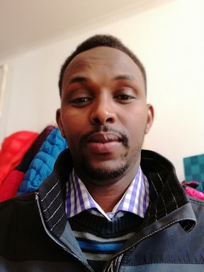 A person wearing a multi-layered outfit with a checked shirt, striped sweater, and dark jacket. The background is a plain wall with clothing items hanging.