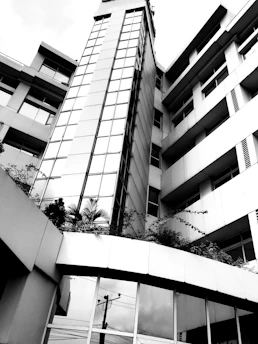 A modern multi-story building with a sleek design features large glass windows and metal paneling. The structure is surrounded by minimal landscaping, including some plants near the base of the building. The architecture is geometric and clean, emphasizing vertical lines.