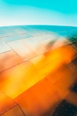 A close-up view of a metal surface composed of large, overlapping panels reflecting vibrant colors. The panels showcase a gradient of hues ranging from orange to blue, possibly due to lighting or reflections. The metal has rivets along its surface, adding texture to the image.