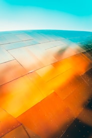 A close-up view of a metal surface composed of large, overlapping panels reflecting vibrant colors. The panels showcase a gradient of hues ranging from orange to blue, possibly due to lighting or reflections. The metal has rivets along its surface, adding texture to the image.