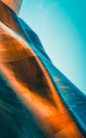 A close-up view of a large, metallic structure with a flowing, curved design. The surface features a mix of warm orange tones and cool blue shades, creating an iridescent effect. The reflective material captures and blends light, producing smooth transitions between colors.