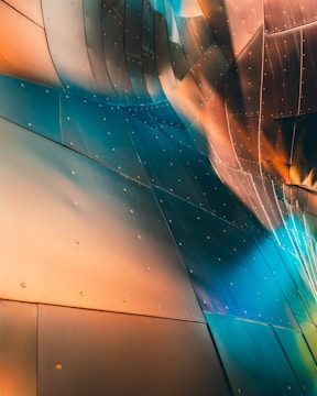 A close-up view of a metallic surface with a smooth, curving design. The material reflects light in a gradient of colors including copper, blue, and teal, creating an abstract, artistic appearance. There are visible rivets evenly spaced across the panels, adding texture to the surface.