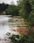 A serene lakeside scene with calm water bordered by lush greenery and tall trees. A small dock hosts a canoe on the right, and a charming house with a chimney is visible across the lake surrounded by more trees.