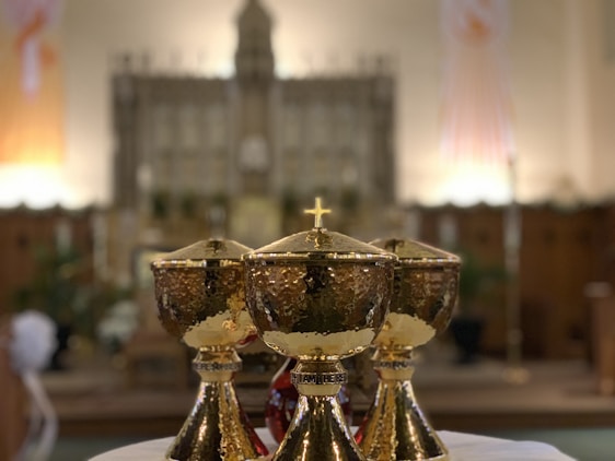 Three golden chalices are prominently placed in the foreground on a white cloth. They have a textured surface with a small cross on the lid. The background is blurred, featuring what appears to be the interior of a church or cathedral, with a large altar and ornate decorations.