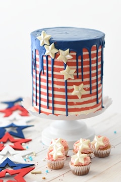 A large round cake decorated with horizontal red and white stripes and blue icing dripping from the top. The cake is adorned with white star-shaped decorations. Beside it, there are small cupcakes topped with similar star-shaped decorations. The setup includes scattered red, white, and blue star cut-outs and sprinkles on a light-colored wooden surface.
