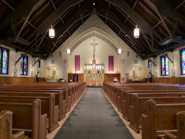 A spacious church interior with wooden pews arranged in rows leading to an ornate altar at the front. High arched ceilings feature exposed wooden beams and hanging lanterns, while colorful stained glass windows line the walls.