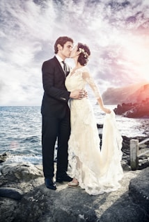 A couple wearing formal attire stands on rocky terrain by the sea, sharing a kiss. The sky is partly cloudy with a touch of sunlight creating a soft glow. The woman is in a lace wedding dress and the man is in a black suit.