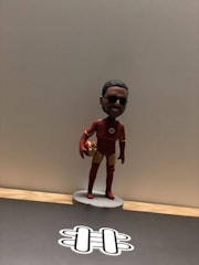 A small figurine of a person wearing a superhero costume, with red and gold colors, stands on a simple circular base. The figurine is holding a ball in one hand and is positioned against a plain backdrop. The figure has sunglasses and a smiling expression.