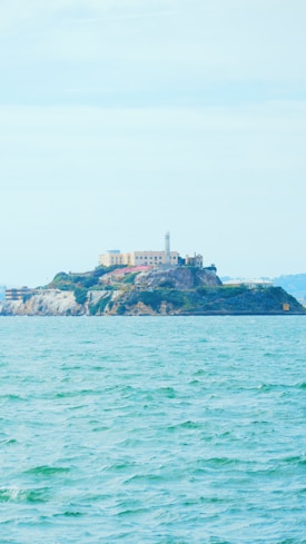 An island with a large building and lighthouse is surrounded by a body of water. Vegetation covers parts of the rocky landscape. The water appears calm with gentle waves.