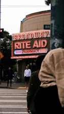 A street scene featuring a neon sign for 'Broadway Rite Aid Pharmacy' with several people standing on the sidewalk. The sign is brightly lit with red and blue colors, mounted on a building that appears to be on a corner. Trees are visible in the background, and there are traffic lights and crosswalk lines in the foreground.