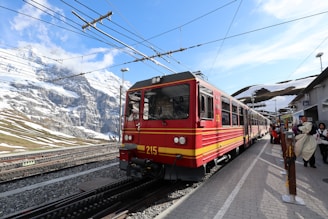 A bright red train is stopped at a platform in a mountainous area. There are snowy peaks in the background, with clear blue skies and some clouds. Passengers are visible on the platform, some carrying bags and wearing coats, indicating a cold climate.