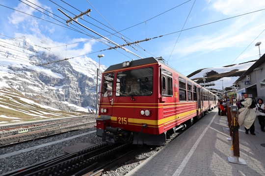 A bright red train is stopped at a platform in a mountainous area. There are snowy peaks in the background, with clear blue skies and some clouds. Passengers are visible on the platform, some carrying bags and wearing coats, indicating a cold climate.