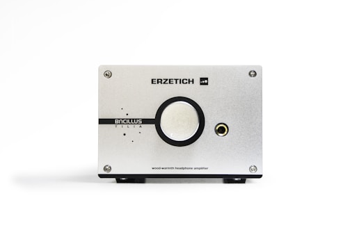 A sleek, minimalist headphone amplifier with a silver metallic finish. It features the brand name 'Erzetich' at the top and 'Bacillus Tilia' below a central large knob. A headphone jack is visible on the right side. The device is sturdy, with screws at each corner and a clean, industrial design aesthetic.