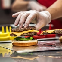 A gloved hand carefully assembling a cheeseburger, placing a layer of cheese and a beef patty onto a bun with sliced green bell peppers. The background includes blurred elements of a kitchen setting with condiments visible.