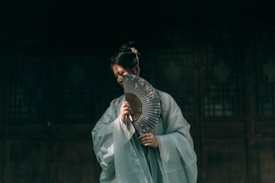 A person dressed in traditional attire is holding an intricately designed fan, partially obscuring their face. The background is dark with wooden paneling, adding a sense of mystery and elegance.