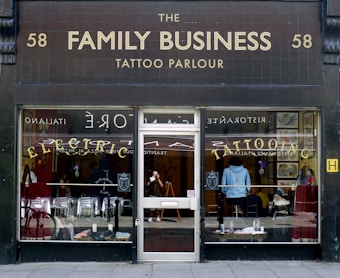 The storefront of a tattoo parlor named The Family Business is visible, with dark tiles displaying gold lettering. Large windows reveal the interior where various tattoo-related decorations and images are visible. The reflection of a street scene can also be seen in the glass.