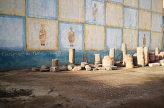 An outdoor scene featuring an ancient-looking mural painted on a wall with repeated images of a person in traditional attire. Several stone columns and fragments are lined up against the wall, suggesting a historical or archaeological setting. The ground appears sandy, and there is a rough, undecorated texture to the scene.