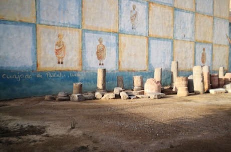 An outdoor scene featuring an ancient-looking mural painted on a wall with repeated images of a person in traditional attire. Several stone columns and fragments are lined up against the wall, suggesting a historical or archaeological setting. The ground appears sandy, and there is a rough, undecorated texture to the scene.