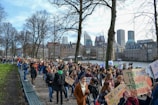 A large group of people is gathered in a peaceful protest, holding various signs with messages related to climate change. The crowd is walking along a path lined with leafless trees, and a cityscape with modern and historic buildings serves as the background.
