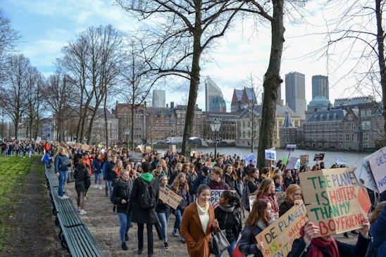 A large group of people is gathered in a peaceful protest, holding various signs with messages related to climate change. The crowd is walking along a path lined with leafless trees, and a cityscape with modern and historic buildings serves as the background.