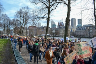 A large group of people is gathered in a peaceful protest, holding various signs with messages related to climate change. The crowd is walking along a path lined with leafless trees, and a cityscape with modern and historic buildings serves as the background.