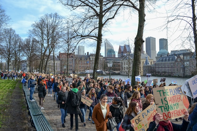 A large group of people is gathered in a peaceful protest, holding various signs with messages related to climate change. The crowd is walking along a path lined with leafless trees, and a cityscape with modern and historic buildings serves as the background.