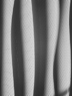 A textured surface with perforated holes arranged in a regular, repeating pattern. The material appears metallic, reflecting light and casting shadows in a rhythmic, wave-like form across the surface.
