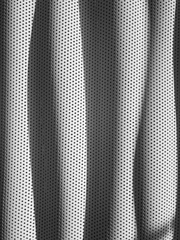 A textured surface with perforated holes arranged in a regular, repeating pattern. The material appears metallic, reflecting light and casting shadows in a rhythmic, wave-like form across the surface.