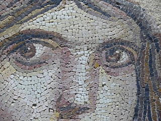 A detailed mosaic artwork depicting a close-up of a face, made with small, multicolored tiles carefully arranged to form distinct features including eyes, nose, and hair. The mosaic demonstrates intricate craftsmanship with shades of beige, brown, and pink.