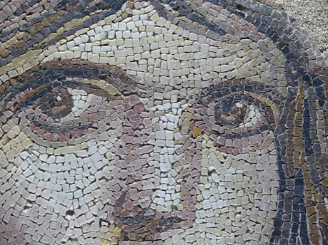 A detailed mosaic artwork depicting a close-up of a face, made with small, multicolored tiles carefully arranged to form distinct features including eyes, nose, and hair. The mosaic demonstrates intricate craftsmanship with shades of beige, brown, and pink.
