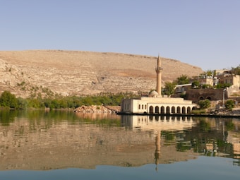 A serene waterfront scene with a historical building featuring arches and a tall minaret, reflecting in the calm waters. The background consists of arid mountains and sparse vegetation, while the foreground includes lush green vegetation along the riverbank.