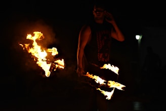 A person is performing a fire dance with flames emanating from sticks. The scene is dimly lit with the bright fire contrasting against the darkness. The performer appears to be skillfully manipulating the fire, creating dynamic shapes.