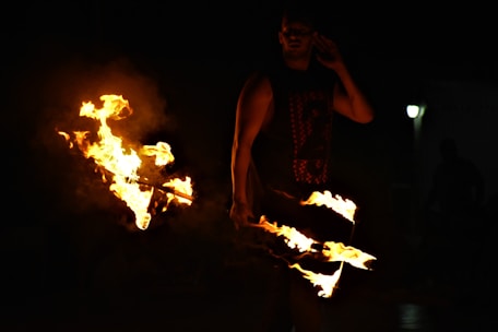 A person is performing a fire dance with flames emanating from sticks. The scene is dimly lit with the bright fire contrasting against the darkness. The performer appears to be skillfully manipulating the fire, creating dynamic shapes.