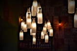 A collection of bottle-shaped pendant lights hanging from the ceiling in a dimly lit interior. The lights glow warmly against a backdrop of eclectic wall art featuring various illustrations and text.