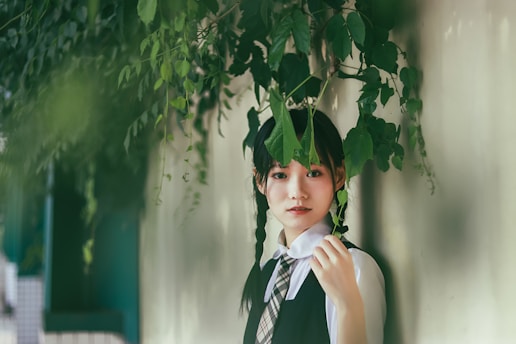 A young person with braided hair stands next to a wall, partially covered with lush green leaves. Their expression is calm and the environment appears serene, with soft lighting enhancing the peaceful atmosphere.
