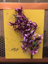 A vibrant yellow canvas is adorned with purple flowers, arranged vertically on the surface. The background is speckled with small purple paint splatters. In the lower portion, there is text written in a decorative script. The canvas is set against a metal mesh backdrop in warm brown tones.
