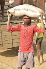A person wearing a red patterned shirt and gray shorts carries a large sack labeled 'Double Bull' on their head. The background shows another person standing near a truck loaded with similar sacks, possibly indicating manual labor in an industrial or construction setting.