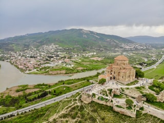 Aerial view of a historic stone church situated on a hilltop. The surrounding landscape features lush greenery, a winding river, and a distant town nestled against rolling hills. The sky is overcast, adding a muted grey tone to the scene. Visitors are visible walking around the church, which is surrounded by ancient ruins.