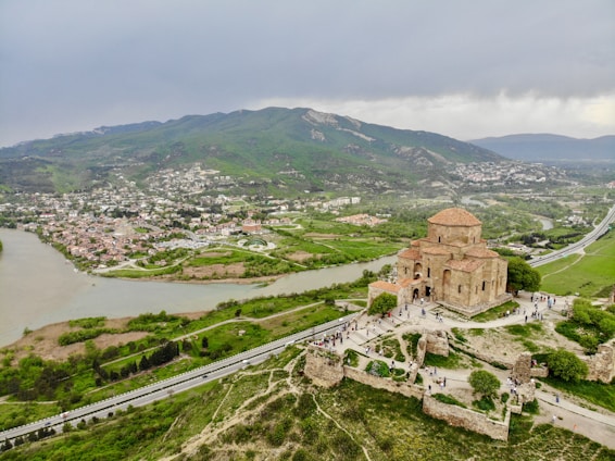 Aerial view of a historic stone church situated on a hilltop. The surrounding landscape features lush greenery, a winding river, and a distant town nestled against rolling hills. The sky is overcast, adding a muted grey tone to the scene. Visitors are visible walking around the church, which is surrounded by ancient ruins.