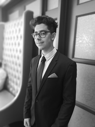 A well-dressed person is standing confidently, wearing a suit with a pocket square. The background includes a tufted chair and decorative wall panels, suggesting a stylish, possibly formal setting.