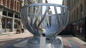 A large, metallic, abstract sculpture resembling a cup is situated in an urban setting. The sculpture features vertical, curved metal strips that create an open, basket-like design. Surrounding the sculpture, there are historic brick and modern glass buildings along with shops displaying Canadian flags. Flower pots and benches are arranged along the pedestrian walkway.