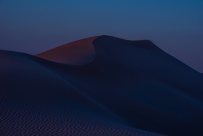 Gently rolling sand dunes are captured under a dimly lit sky, creating a serene and mysterious landscape. The smooth curves of the dunes are highlighted by a subtle gradient of colors as twilight sets in.