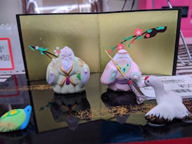 Two ceramic figurines in traditional attire are displayed against a decorative gold backdrop adorned with branches and flowers. A white crane figurine sits to the right, while a small colorful turtle is in the foreground. Price tags are visible in the background.
