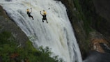 Two people wearing helmets are zip-lining across a large, powerful waterfall. They are suspended high above the ground, amidst a lush, green forested area. The waterfall cascades down steep rocks, creating a dramatic and adventurous setting.