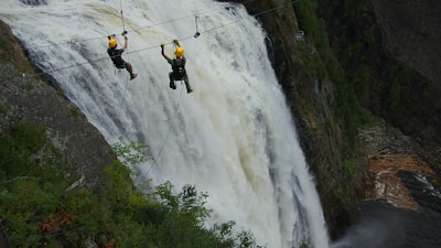 Two people wearing helmets are zip-lining across a large, powerful waterfall. They are suspended high above the ground, amidst a lush, green forested area. The waterfall cascades down steep rocks, creating a dramatic and adventurous setting.