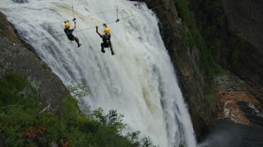 Two people wearing helmets are zip-lining across a large, powerful waterfall. They are suspended high above the ground, amidst a lush, green forested area. The waterfall cascades down steep rocks, creating a dramatic and adventurous setting.