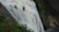 Two people wearing helmets are zip-lining across a large, powerful waterfall. They are suspended high above the ground, amidst a lush, green forested area. The waterfall cascades down steep rocks, creating a dramatic and adventurous setting.