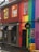 A vibrant building features a rainbow-colored facade with stripes of red, orange, yellow, green, blue, and purple. The upper windows have bright orange and yellow frames. There is a sign for 'Kiki queer bar' above the entrance, and several black awnings cover the windows below. The door is painted in black and a sidewalk with a rainy surface is in front of the building. A wooden box is placed near the entrance.